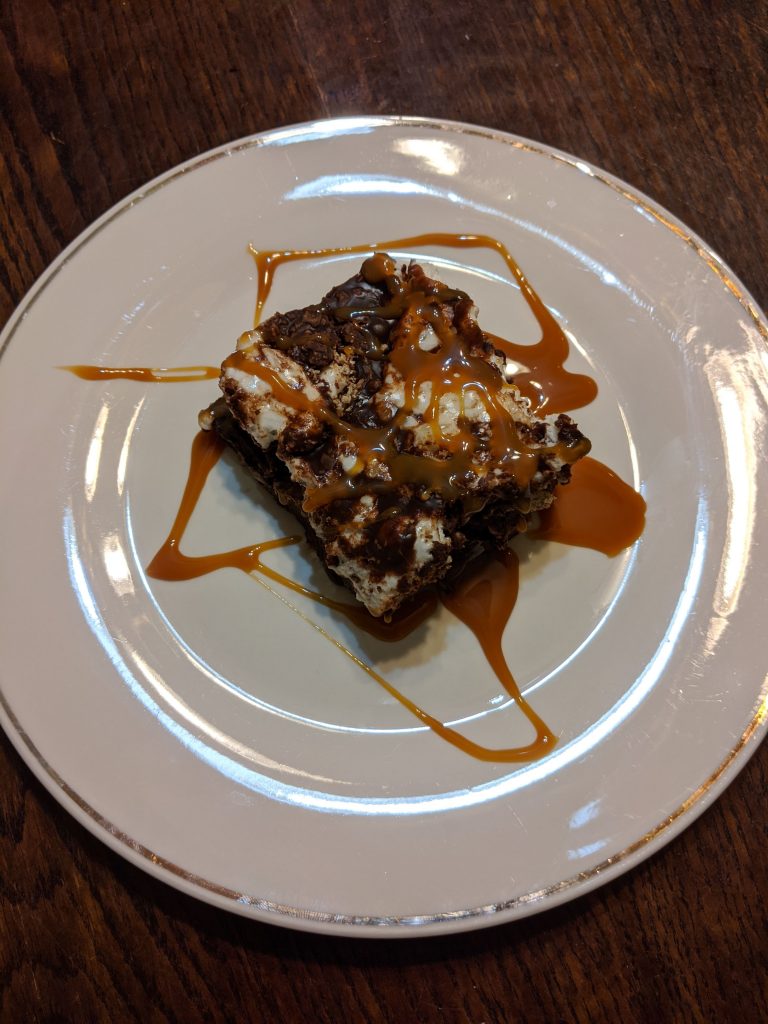 Birds eye view of a loaded ganache brownie topped with marshmallow, chocolate pieces, and caramel syrup on a white dish.