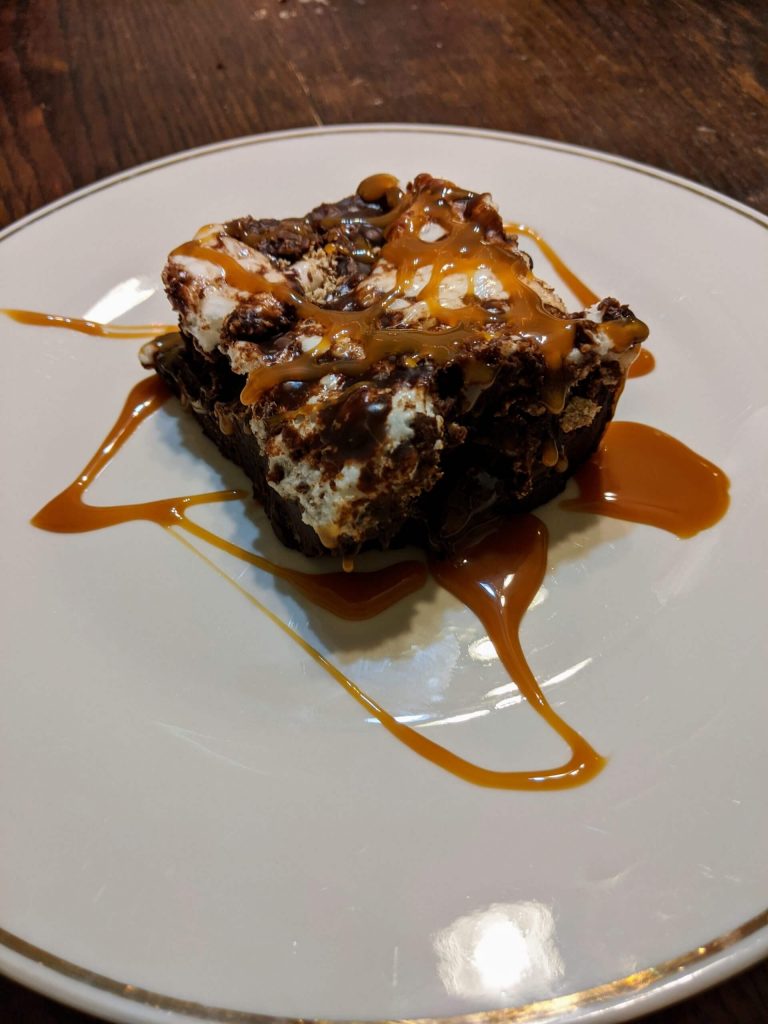 Loaded ganache brownie topped with marshmallow, chocolate pieces, and caramel syrup on a white dish.