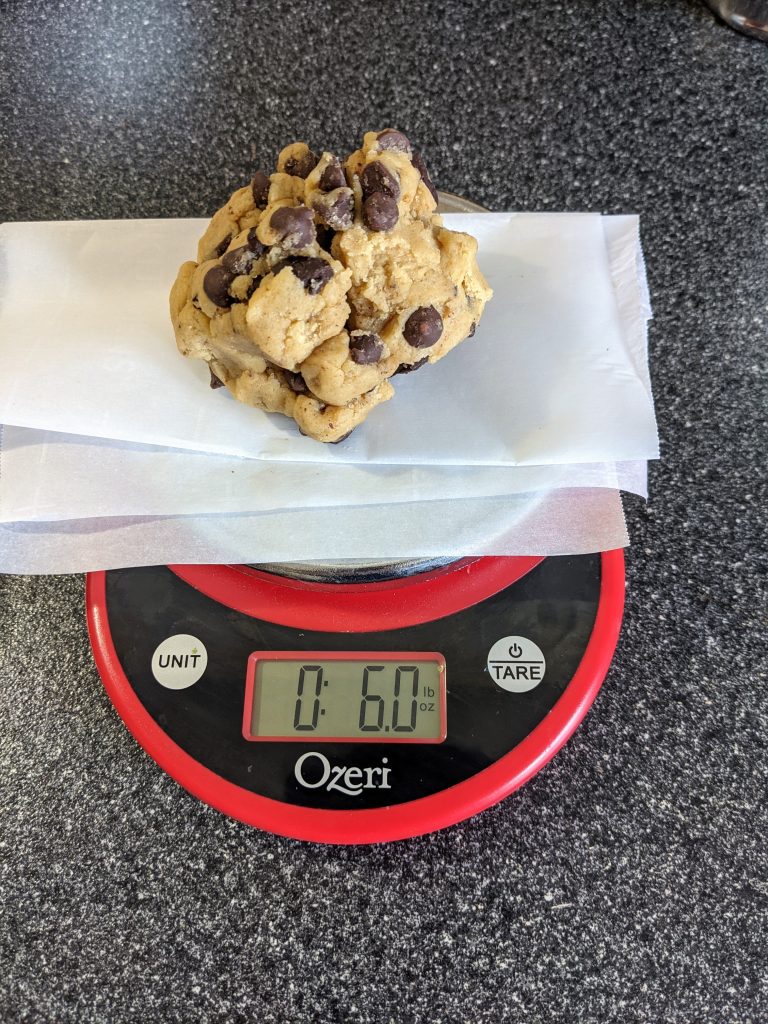 One large chocolate chip cookie dough ball sitting mon a scale that reads 6.0 oz.