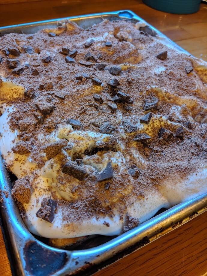 Side view of finished chocolate tres leches covered with Cool Whip, chocolate crumbs and pieces sitting in a baking pan.