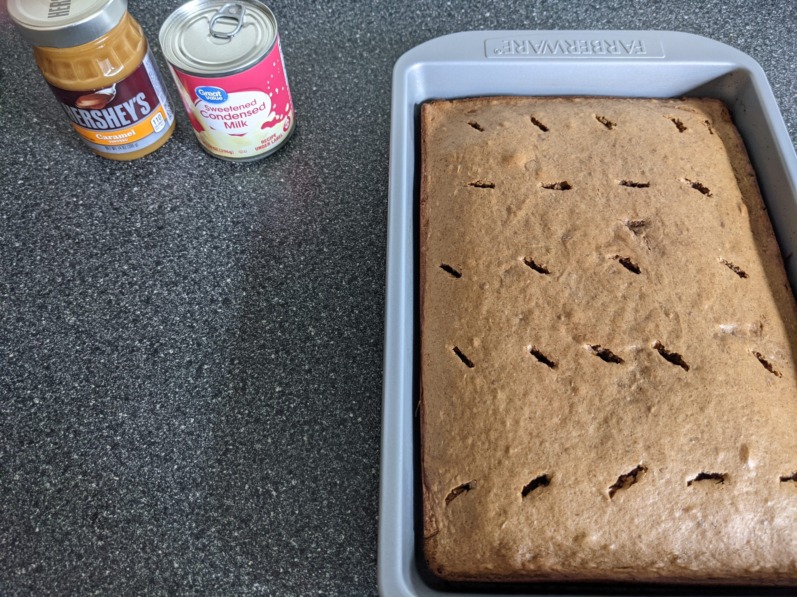 Baked German chocolate cake in pan with holes scattered in it, caramel and sweetened condensed milk can next to it.