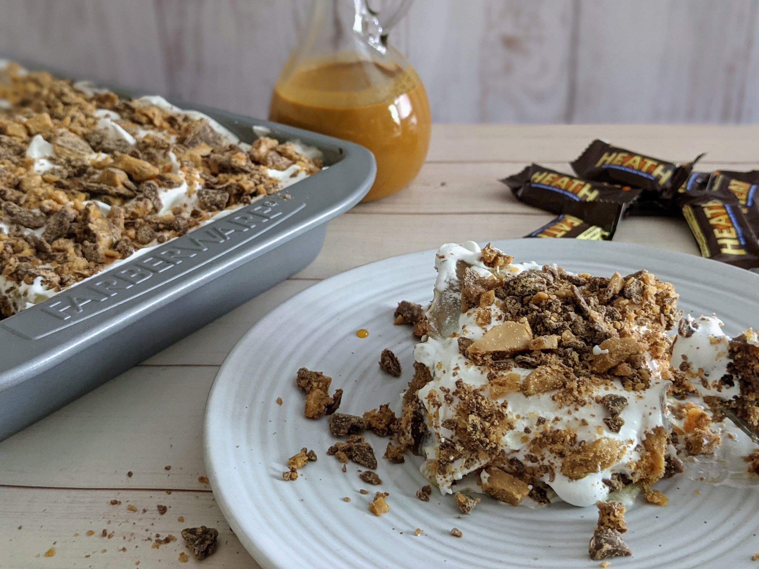 Side eye view of a finished Greggs Heath bar cake in a pan in the corner, heath bar candy and a slice of heath bar cake next to the pan.