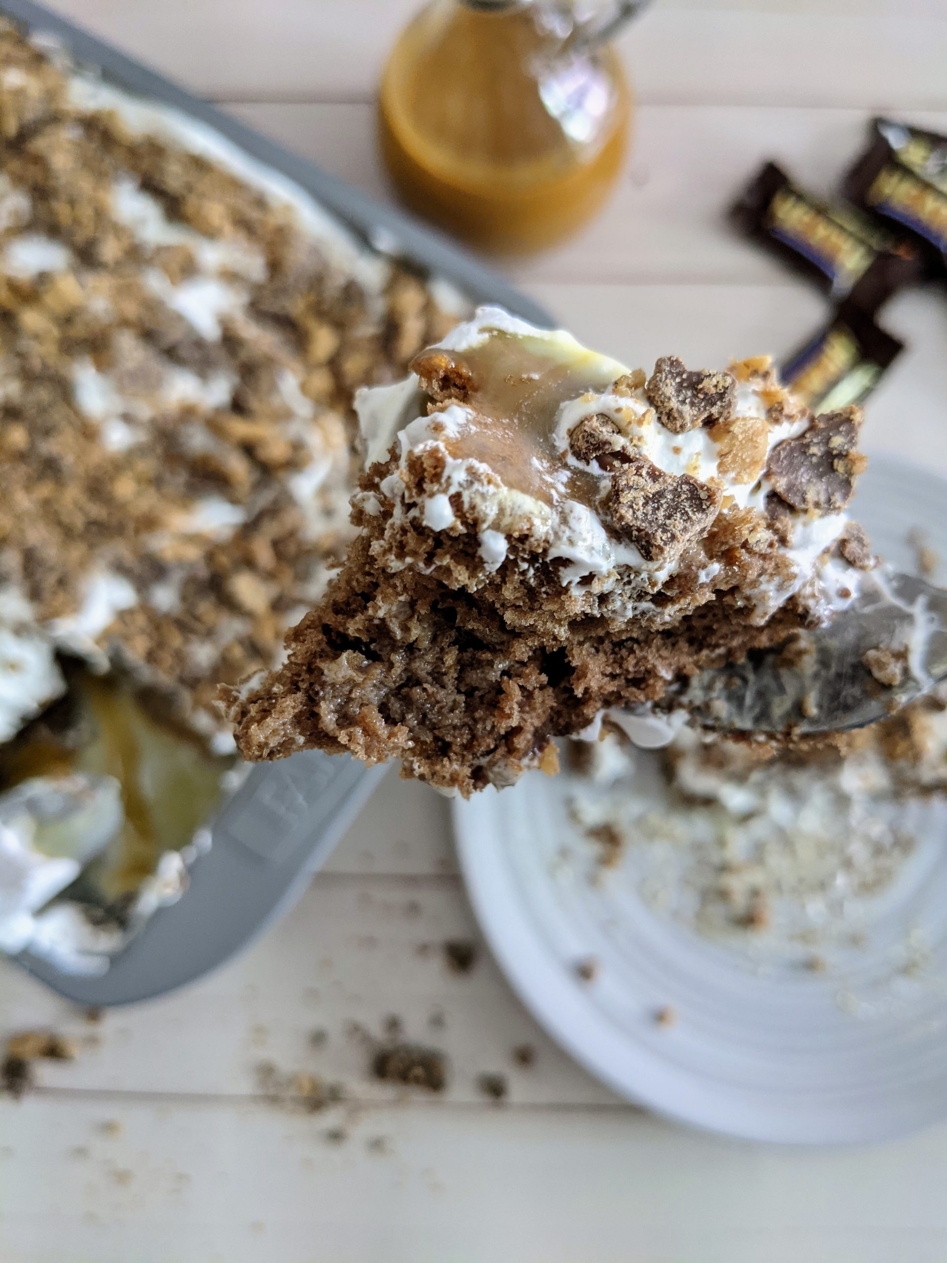 Piece of heath bar cake on a fork with a blurred background of birds eye view of a finished Greggs Heath bar cake in a pan in the corner, heath bar candy and a slice of heath bar cake next to the pan.