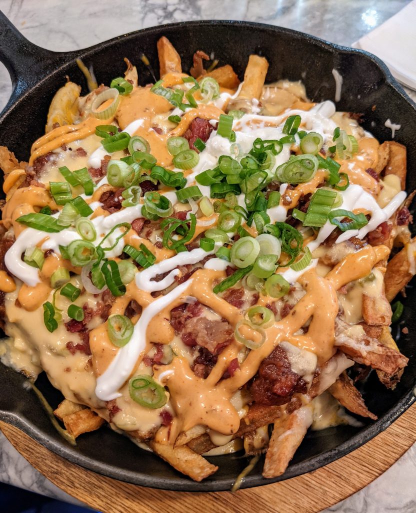 Large black pan of loaded fries with cheese, bacon, and a green type plant seasoning.