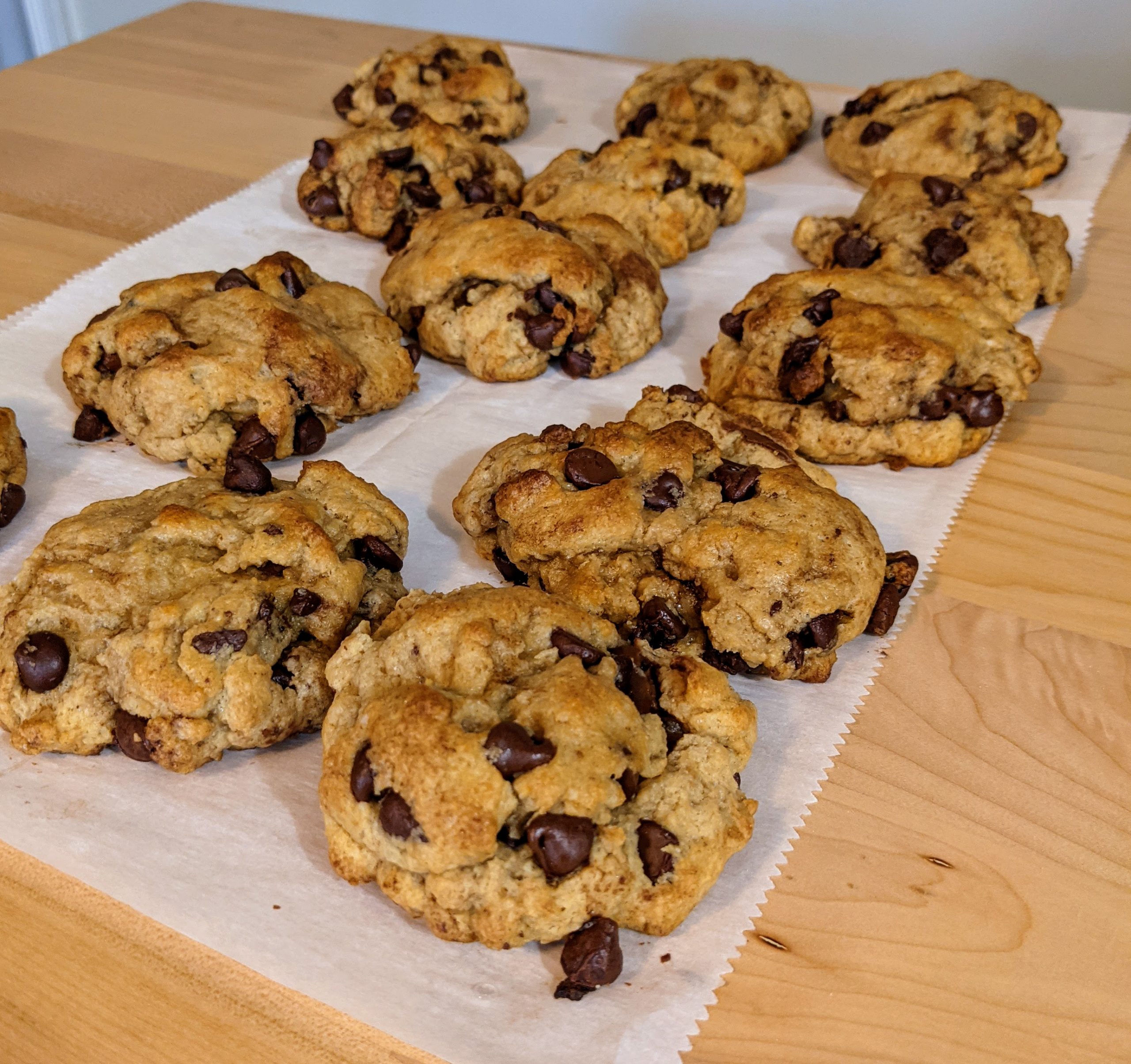 Several finished cookies filled with chocolate chips and Nutella sitting on parchment paper on a wooden table after sitting out for an hour.