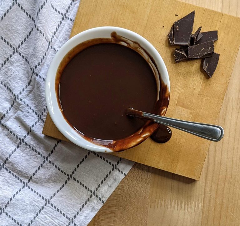Birds eye view of chocolate sauce in a white bowl next to chopped up chocolate pieces on a cutting board near a white and brown napkin.