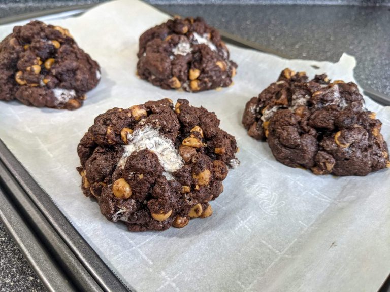 Four large finished dark chocolate cookies with peanut butter chips and oozing marshmallow on a parchment lined baking sheet.