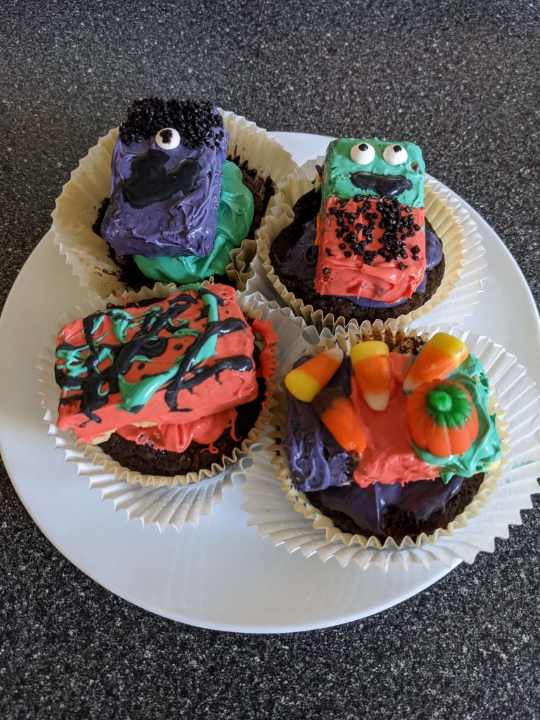 Four chocolate brownies on a white plate each topped with a decorated Halloween Krispy Treat.