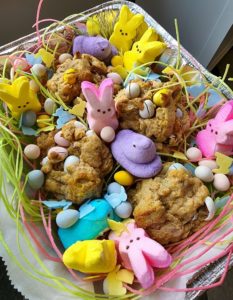 Birds eye view of a few large cookies with Easter egg chocolate inside them sit inside a baking tin with peeps and edible grass.