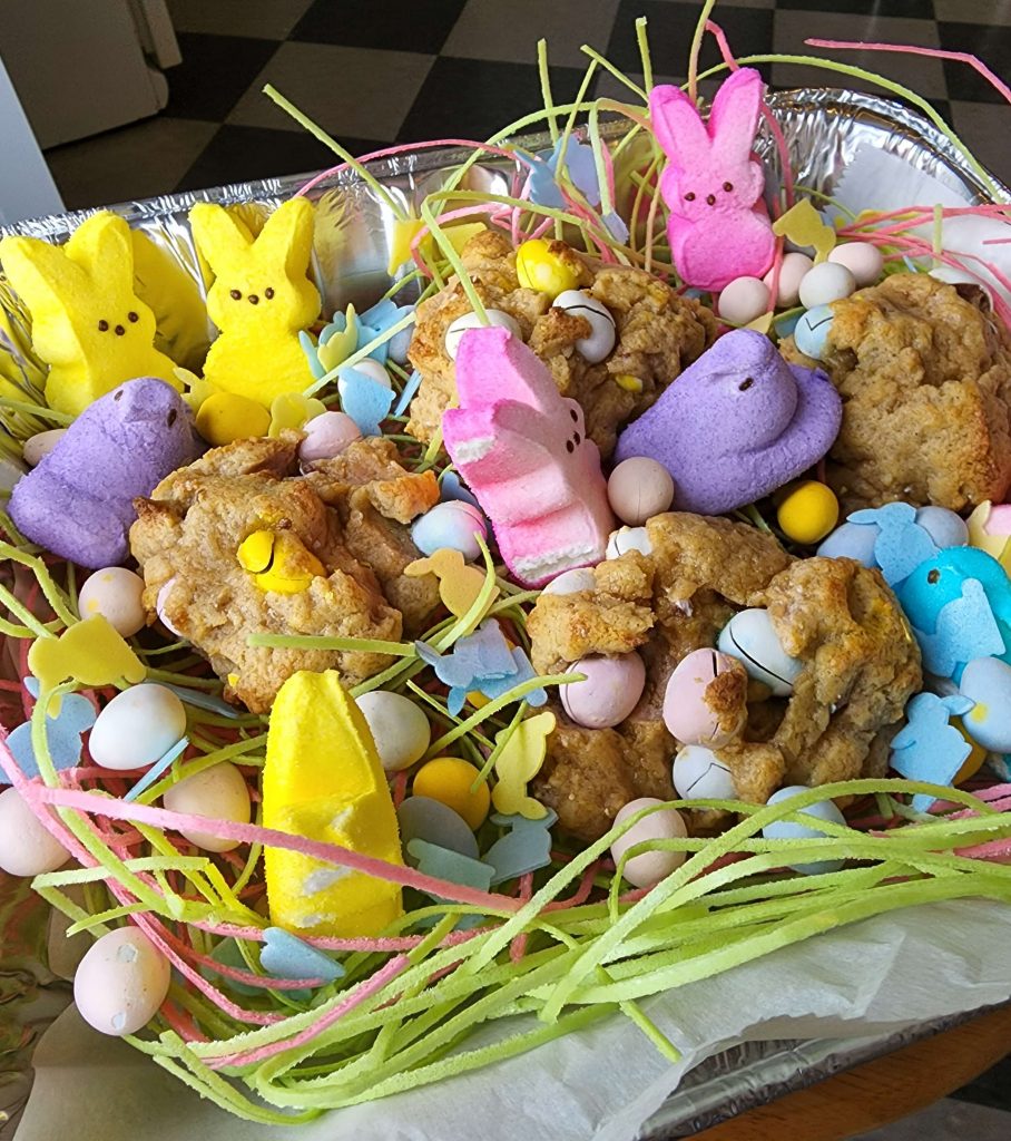 A few large cookies with Easter egg chocolate inside them sit inside a baking tin with peeps and edible grass.