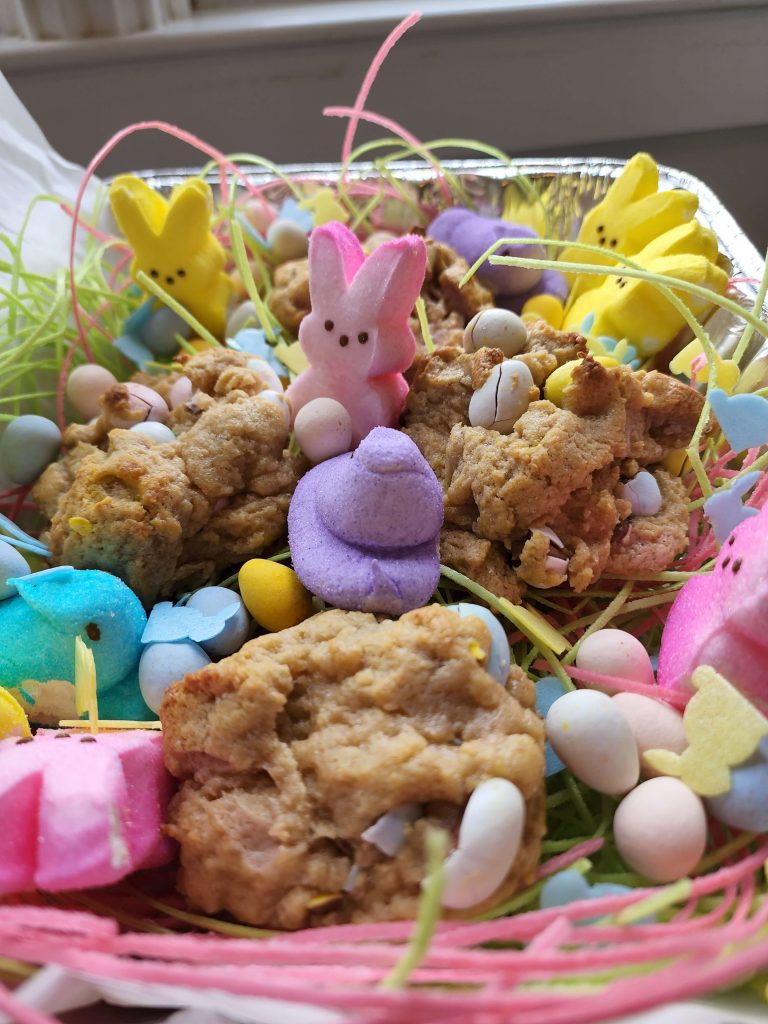 A few large cookies with Easter egg chocolate inside them sit inside a baking tin with peeps and edible grass.