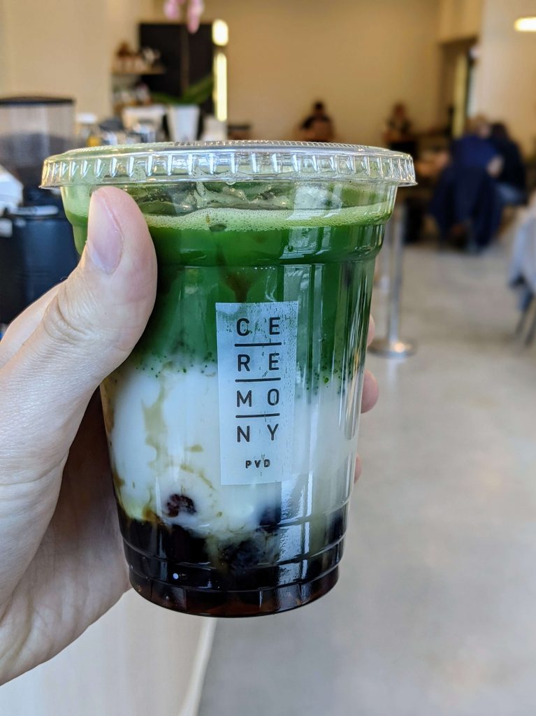 Holding a three layer bubble tea, top is green matcha, middle is milky mochi, and the bottom is boba pearls. 