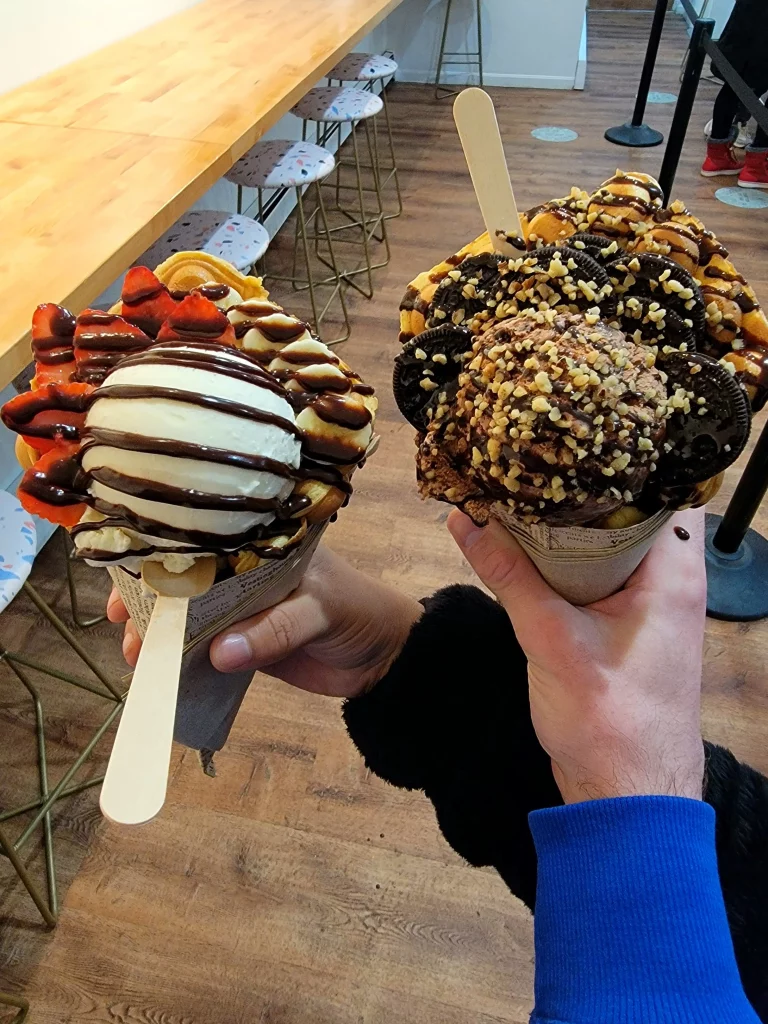 Two hong kong bubble waffle ice creams being held. One with chocolate ice cream and nut and Oreo toppings, the other with vanilla ice cream, chocolate drizzle, and strawberries.