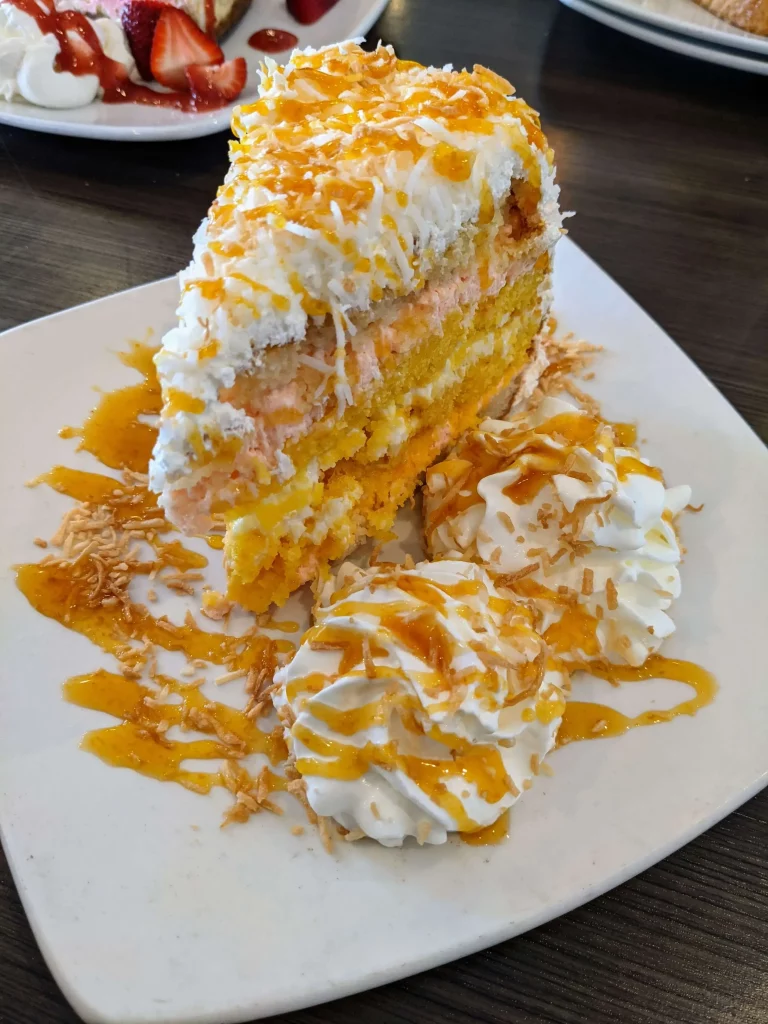 Close up of a large slice of yellow-white looking cake with white frosting and topping and lined with fruit pieces and whipped cream.