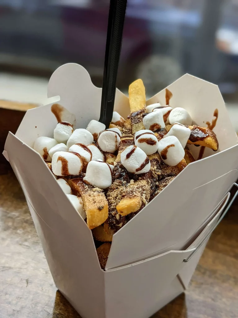 French fries in a Chinese takeout container topped with chocolate syrup, cracker crumbs, and mini marshmallows.