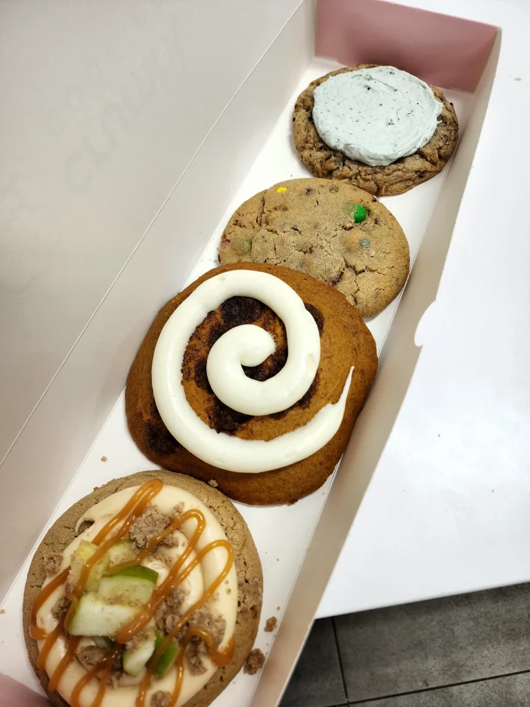 Four Crumbl Cookies up close lined up in a pink and white box. Caramel Apple, Pumpkin Roll, M&M, and Aggie Blue Mint.