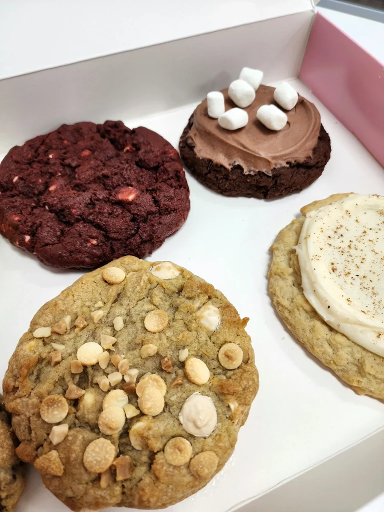 Four Crumbl Cookies up close in a pink and white box. Red Velvet, Frozen Hot Chocolate, Eggnog, and Macadamia Nut.