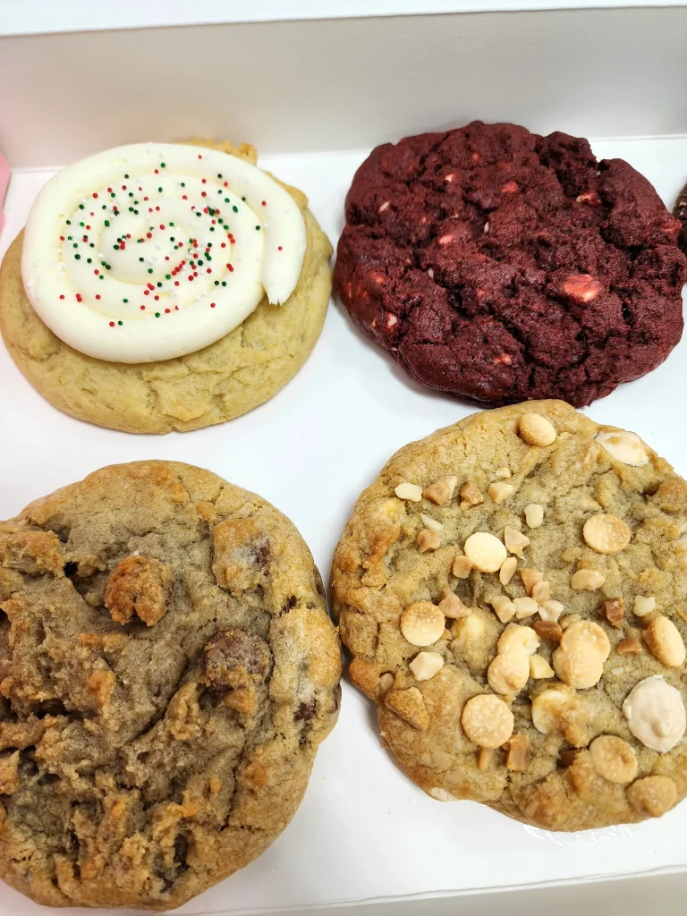 Four Crumbl Cookies up close in a pink and white box. Red Velvet, Birthday Cake, Chocolate Chip, and Macadamia Nut.