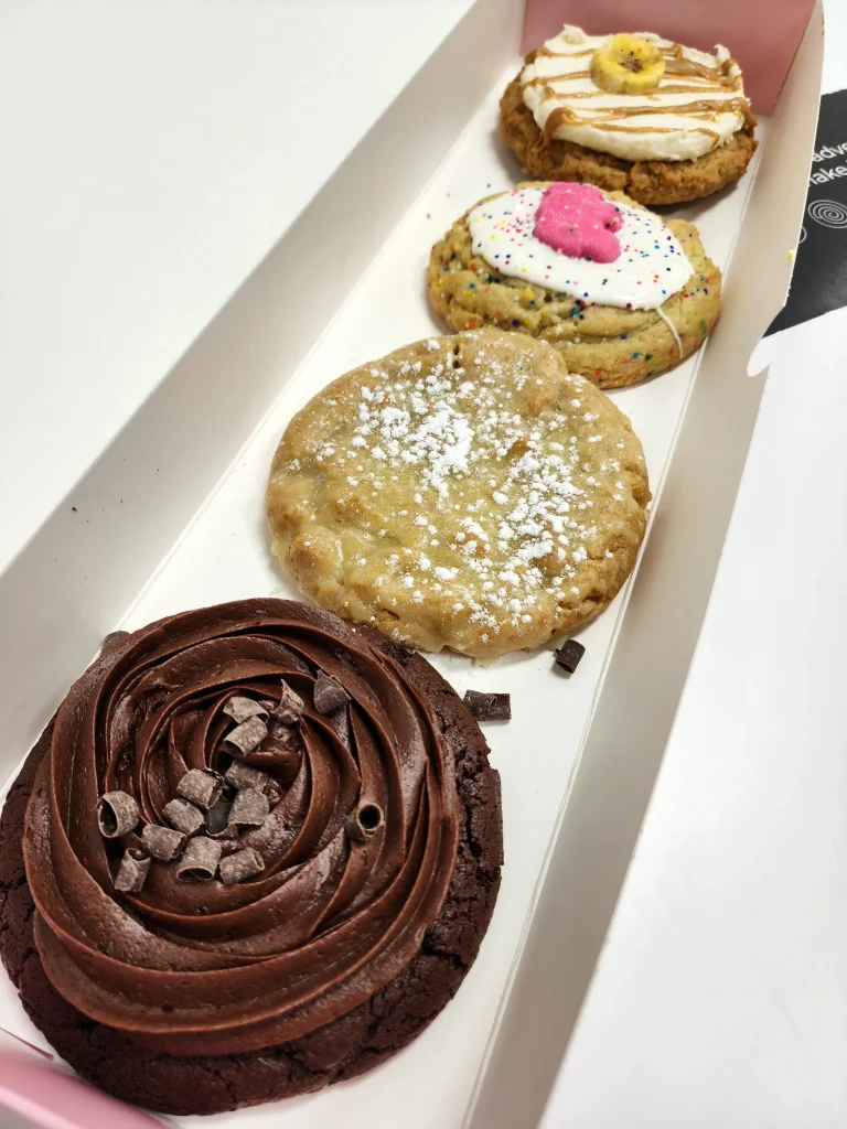 Four Crumbl Cookies up close lined up in a pink and white box. Sugar ft. Mother's Circus Animal, Chocolate Cake, Kentucky Butter Cake, and Peanut Butter Banana.