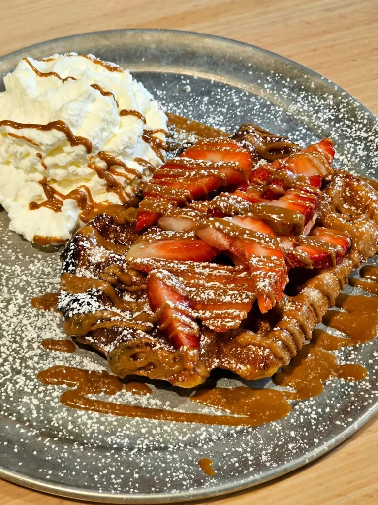 Fresh waffle shop Liege waffle topped with nut butter, strawberry, and powdered sugar, with a side of whipped cream.