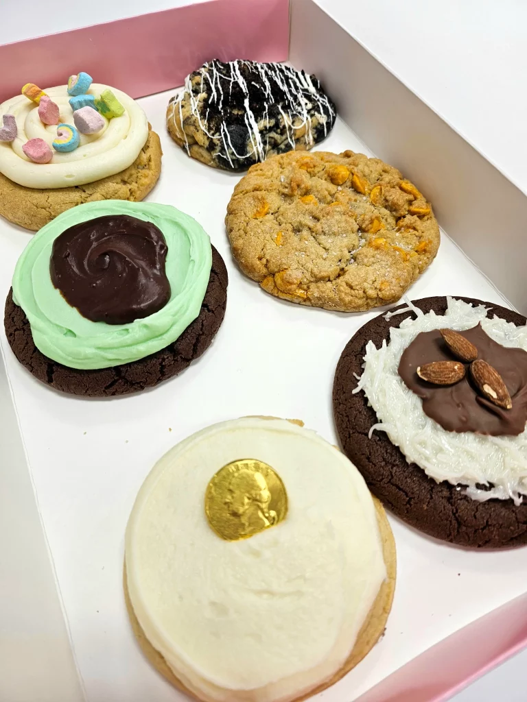 Six Crumbl Cookies up close in a pink and white box. Cookies & Cream, Mallow Creme, Sugar (Gold Coin), Mint Brownie, Butterscotch Chip, Almond Coconut Fudge.
