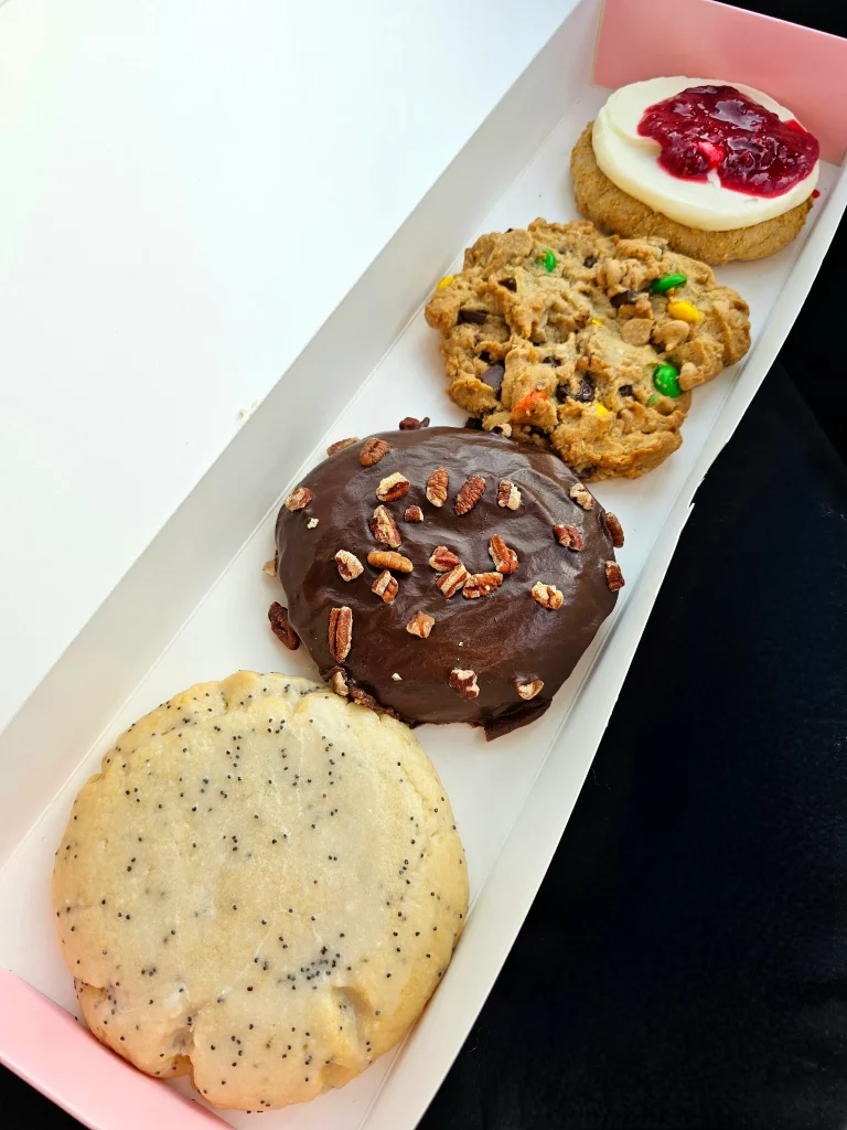 Four Crumbl Cookies up close lined up in a pink and white box. Lemon Poppy Seed, Texas Sheet Cake, Raspberry Cheesecake, and Monster.