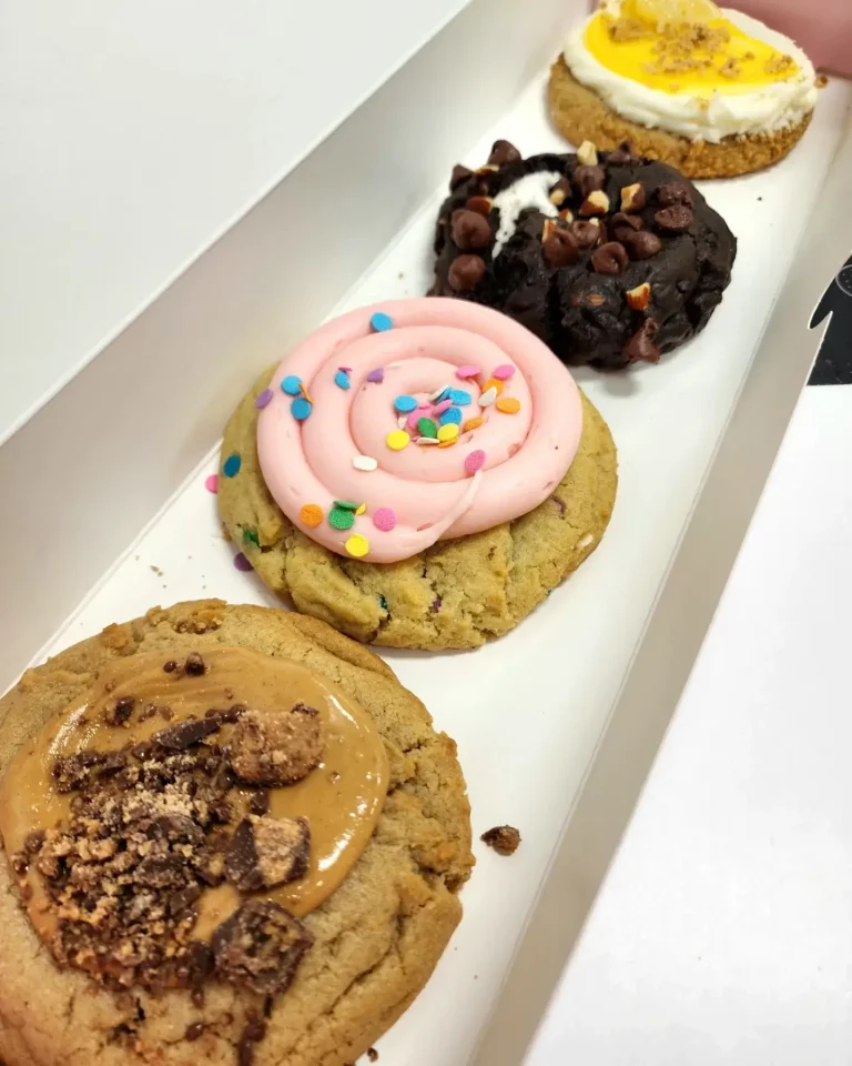 Four Crumbl Cookies up close lined up in a pink and white box. Ultimate Peanut Butter, Confetti Cake, Lemon Cheesecake, and Rocky Road.