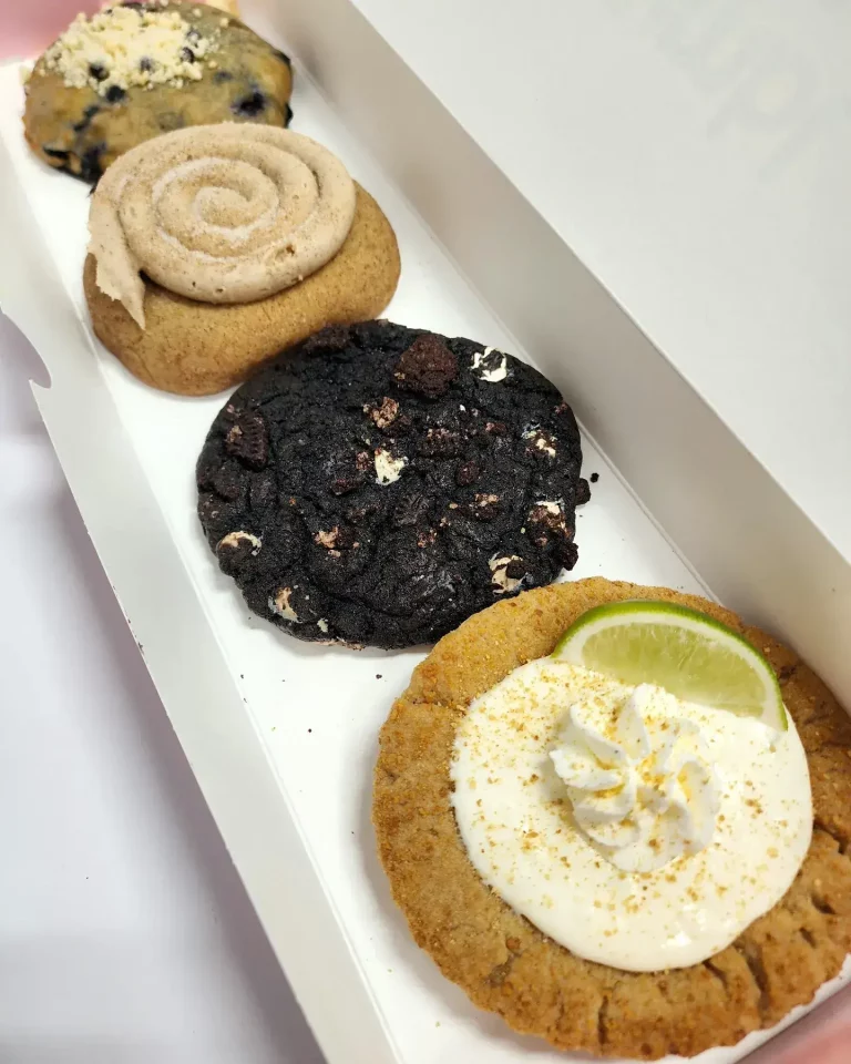 Four Crumbl Cookies up close lined up in a pink and white box. Blueberry Muffin, Keylime Pie, Chocolate Cookies & Cream, and Churro.