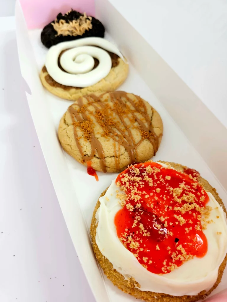 Four Crumbl Cookies up close lined up in a pink and white box. Strawberry Cheesecake, Cookie Butter Lava ft. Biscoff, Cinnamon Roll, and Chocolate Peanut Butter ft. Oreo.