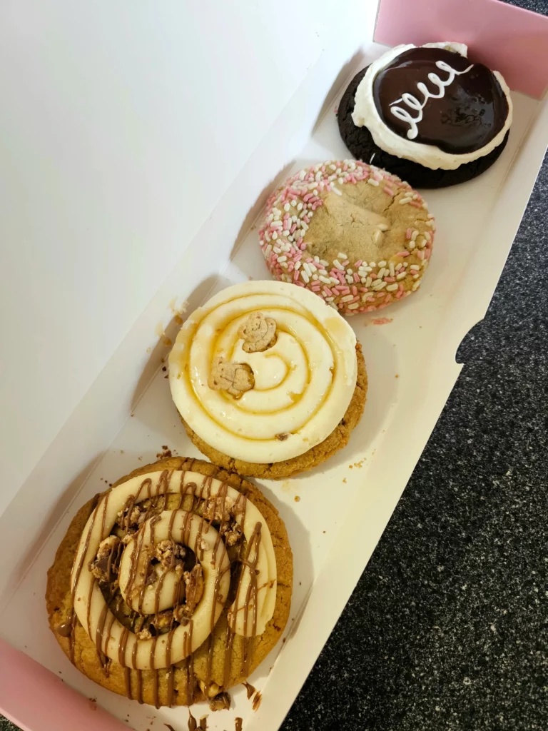 Four Crumbl Cookies up close lined up in a pink and white box. Honey Cake ft. Teddy Grahams, Chocolate Mallow Cupcake, Cake Batter Blondie, and Peanut Butter ft. Snickers