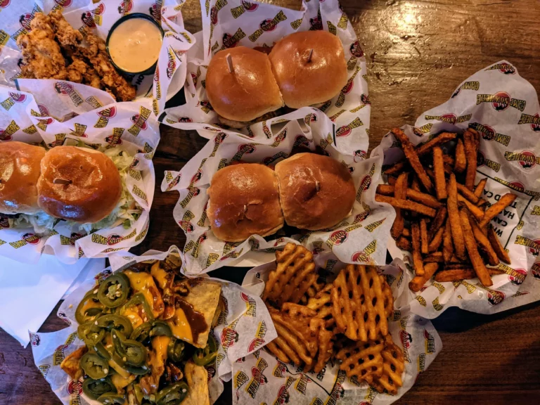 A large variety of food on a wooden table at Camp Nowhere. Includes sliders, waffle fries, French fries, nachos, and more.