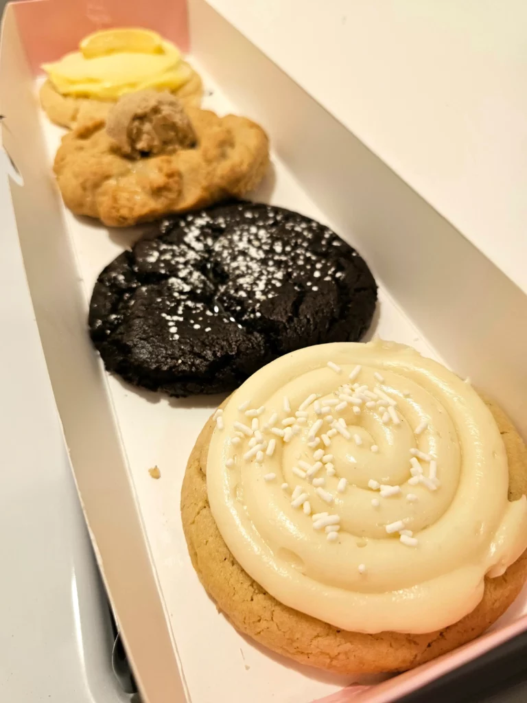 Four Crumbl Cookies up close lined up in a pink and white box. Molten Lava, Cinnamon Frybread, Nilla Bean Cupcake, and Lemonade.