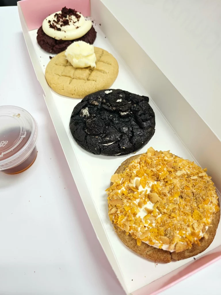 Four Crumbl Cookies up close lined up in a pink and white box. Chocolate Cookies & Cream, Red Velvet Cupcake, Waffle with syrup, and Fried Ice Cream.