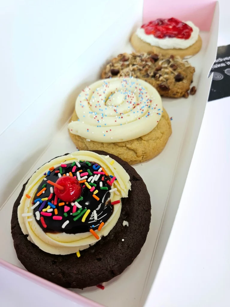 Four Crumbl Cookies up close lined up in a pink and white box. Brownie Sundae, Birthday Cake, Strawberry Shortcake, & Cowboy Cookie.