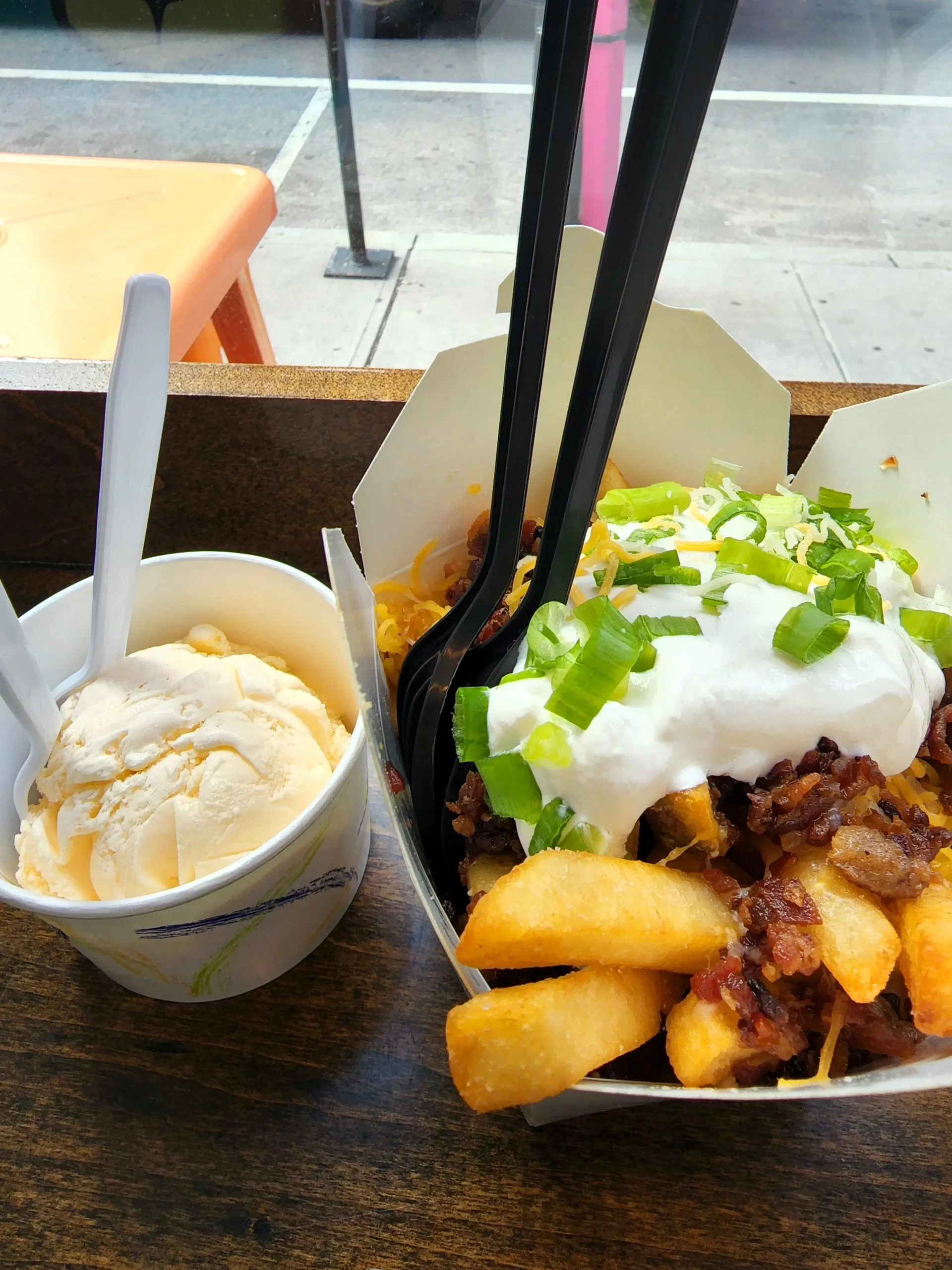 Loaded french fries topped with sour cream, bacon and green shavings with a side of ice cream on a counter.