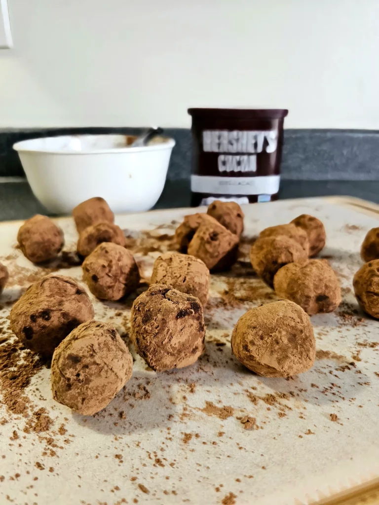 Several Marzipankartoffeln (Marzipan Potato or Cocoa Dusted Marzipan) on a parchment lined wooden board with Hershey's Cocoa powder and a bowl in the background.