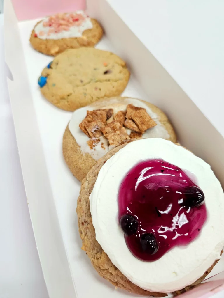 Four Crumbl Cookies up close lined up in a pink and white box. Strawberry Crumb Cake, Original Ft. Peanut M&M's, Berries & Cream, and Cinnamon Crunch.