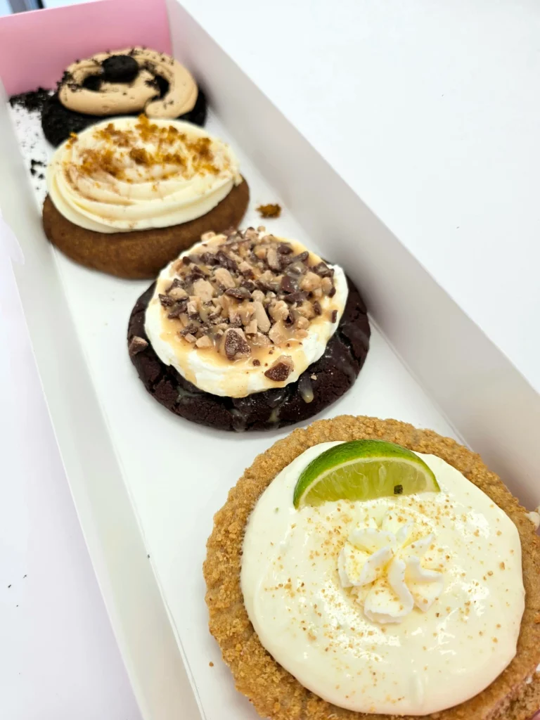 Four Crumbl Cookies up close lined up in a pink and white box. Chocolate Peanut Butter ft. Oreo, Caramel Pumpkin Cake, Key Lime Pie, and Chocolate Toffee Cake.