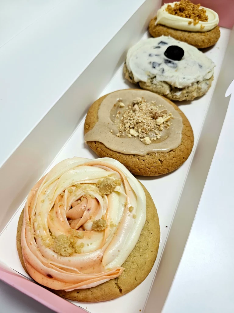 Four Crumbl Cookies up close lined up in a pink and white box. Guava Cake, New York Cheesecake, Cookies n Creme Milkshake ft. Oreo, and Brown Sugar Cinnamon ft. Pop-Tarts.