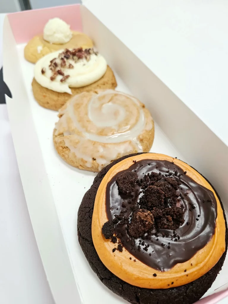 Four Crumbl Cookies up close lined up in a pink and white box. Pumpkin Brownie, Carrot Cake, Buttermilk Pancake, & Honey Bun.