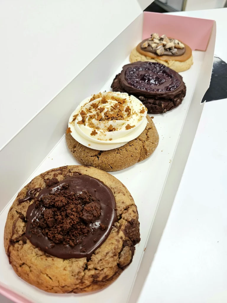 Four Crumbl Cookies up close lined up in a pink and white box. Chocolate Raspberry Truffle, Chocolate Crumb Cake, Gingerbread, & Caramel Shortbread Ft. Twix.