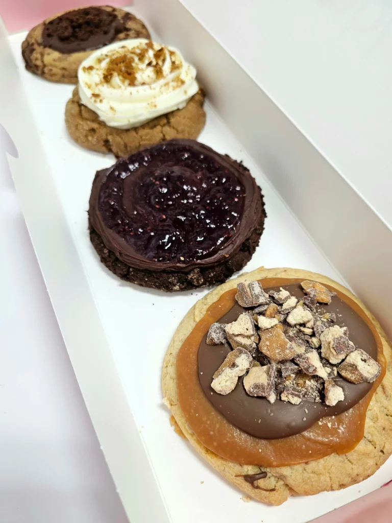 Four Crumbl Cookies up close lined up in a pink and white box. Chocolate Raspberry Truffle, Chocolate Crumb Cake, Gingerbread, & Caramel Shortbread Ft. Twix.
