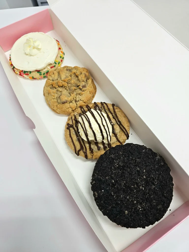 Four Crumbl Cookies up close lined up in a pink and white box. Skillet Cookie, Mallow Sandwich ft. Oreo, Milk Chocolate Chip, and Confetti Milkshake.