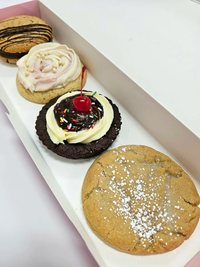 Four Crumbl Cookies up close lined up in a pink and white box. Kentucky Butter Cake, Brownie Sundae, Wedding Cake, Graham Cracker Fudge Sandwich.