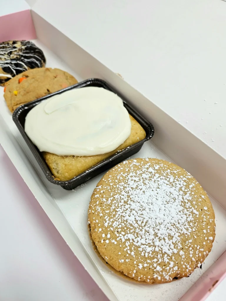 Four Crumbl Cookies up close lined up in a pink and white box. Cookies & Cream, Peanut Butter ft. Reese's Pieces, Cinnamon Square, Cannoli.