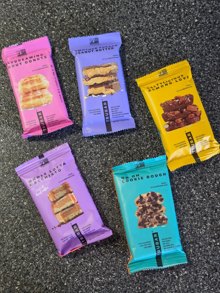 TRUBAR plant-based protein bars, including flavors Daydreaming About Donuts, Smother Fudger Peanut Butter, Saltylicious Almond Love, Whole Lotta Macchiato, and Oh Oh Cookie Dough.