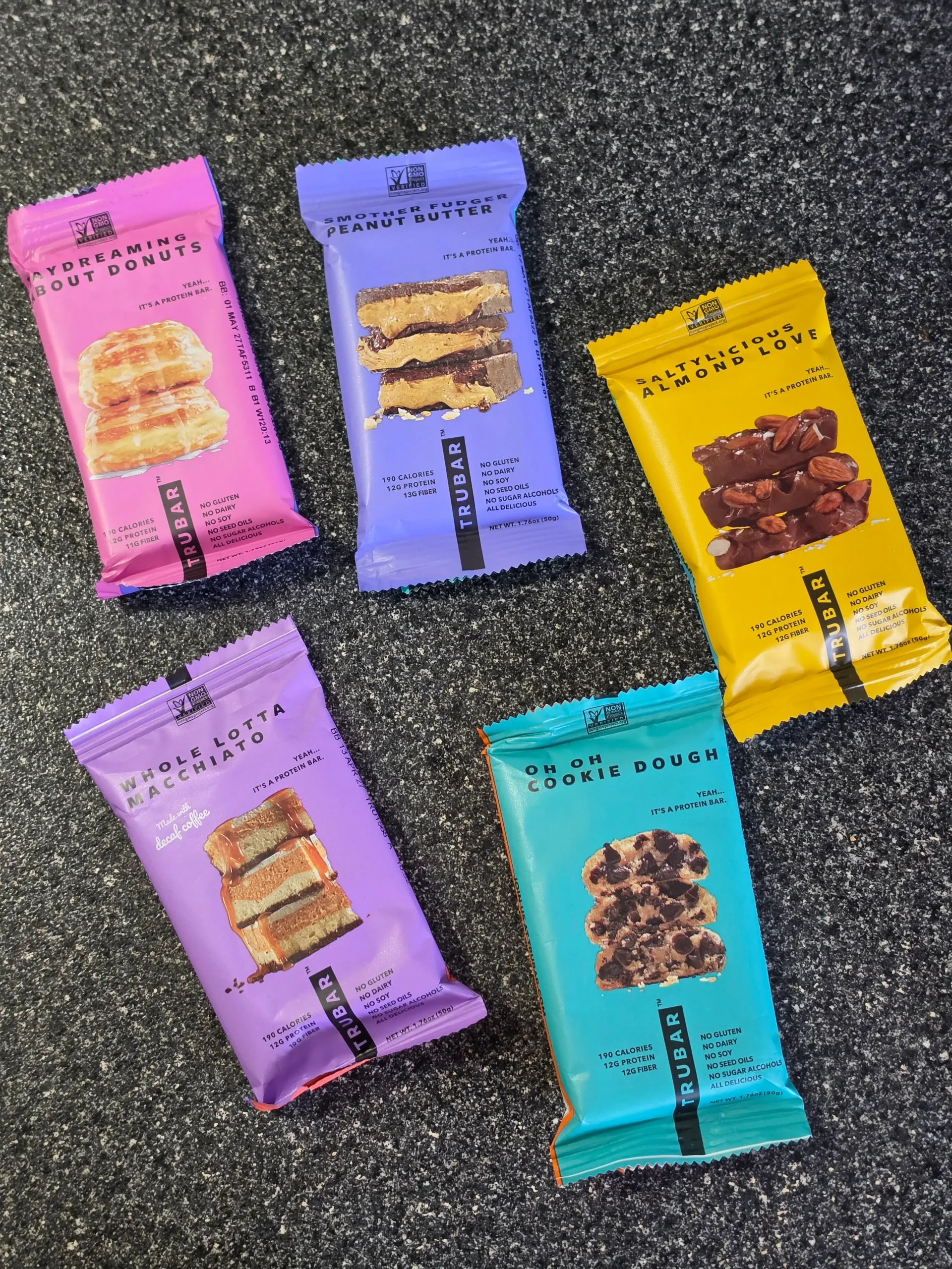 TRUBAR plant-based protein bars, including flavors Daydreaming About Donuts, Smother Fudger Peanut Butter, Saltylicious Almond Love, Whole Lotta Macchiato, and Oh Oh Cookie Dough.
